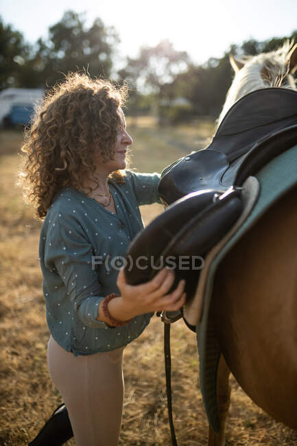 Side view of middle aged female with curly hair putting saddle on back of mare on farm in sunlight — Stock Photo