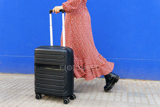 Vista laterale della femmina irriconoscibile ritagliata in lungo abito rosso in piedi con bagagli sulla strada contro il muro blu durante il giorno — Foto stock