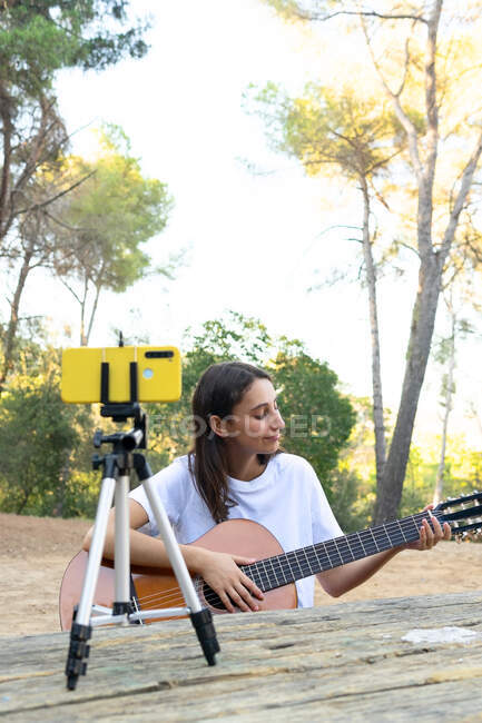 Allegro teen blogger femminile che suona la chitarra acustica mentre registra video sul cellulare su treppiede nel parco — Foto stock