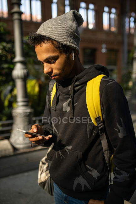 African American male hipster in street style clothes and wireless earphones listening to music while standing in city street and messaging on mobile phone — Stock Photo
