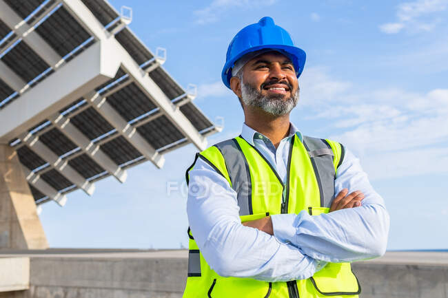 Low angle of delighted ethnic male inspector in vest with protective helmet crossing arms standing against modern solar power station — Stock Photo