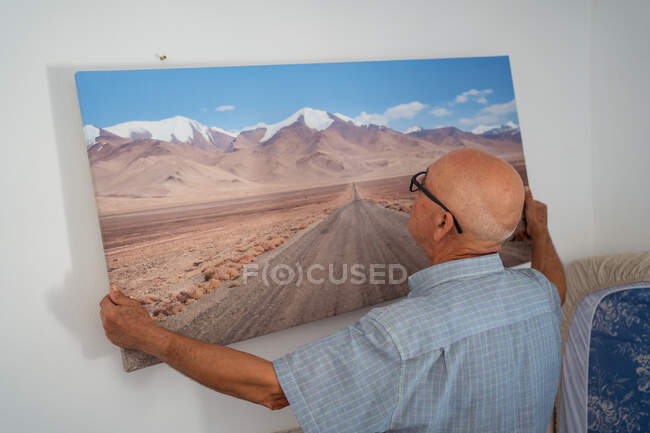 Back view of senior bald male in eyeglasses and checkered shirt hanging artwork of road and mountains on wall at home — Stock Photo
