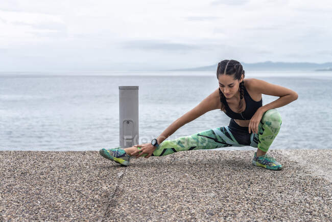 Atleta feminina focada fazendo exercício de lunge lateral e alongamento das pernas enquanto se aquece durante o treinamento em aterro perto do mar — Fotografia de Stock