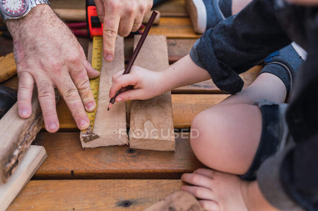 Recadrer papa méconnaissable mesure pièce en bois avec du ruban adhésif contre l'enfant avec crayon assis sur la promenade — Photo de stock