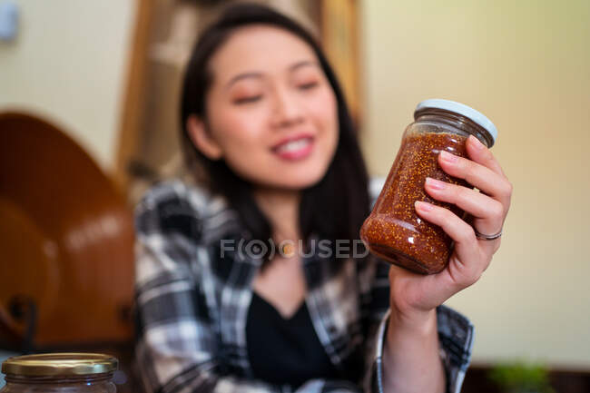 Jeune femme ethnique avec pot en verre de délicieuse marmelade de figue sur table à la maison sur fond flou — Photo de stock