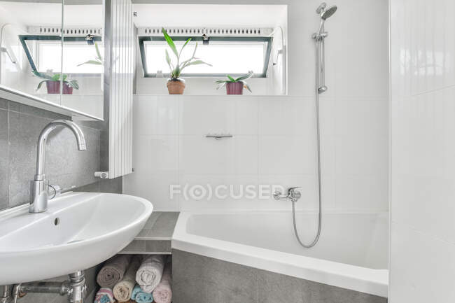Interior de un elegante cuarto de baño contemporáneo con ducha debajo de la ventana y espejo colgando por encima del lavabo cerca de toallas - foto de stock