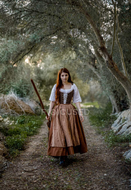 Serious witch in dress standing with a broom on road in woods and looking at camera — Stock Photo