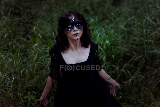Зверху містична відьма в довгій чорній сукні і з пофарбованим обличчям, що стоїть в темному похмурому лісі — стокове фото