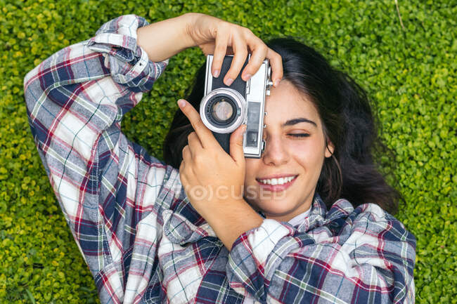 Vista dall'alto della donna sorridente con occhio chiuso in camicia a scacchi che scatta foto sulla fotocamera invecchiata mentre giace sul prato — Foto stock