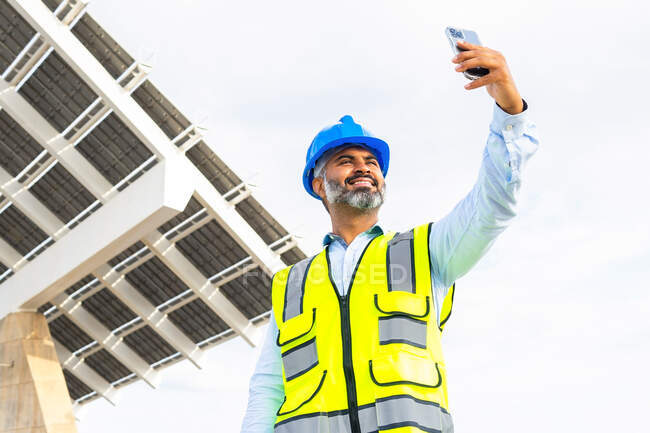 Low angle of cheerful mature ethnic male supervisor in helmet and waistcoat taking self portrait on cellphone against modern construction in sunlight — Stock Photo
