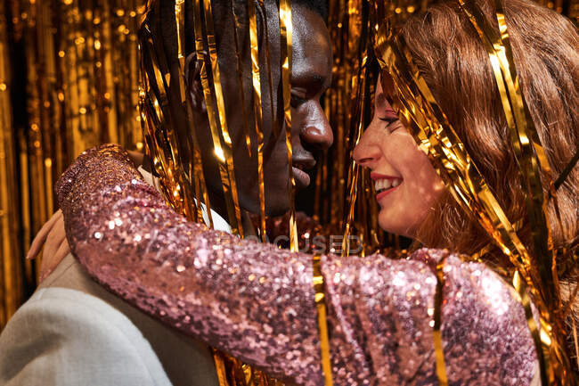 Vista lateral del contenido de la mujer joven en ropa brillante abrazando a la pareja masculina afroamericana mientras se miran durante la fiesta - foto de stock