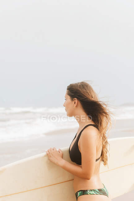 Jeune athlète réfléchie en maillot de bain aux cheveux volants et planche de surf regardant loin sur la côte océanique — Photo de stock