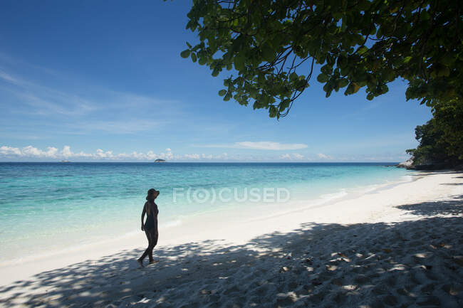 Side view of ethnic female tourist in swimsuit and straw hat walking on sand during trip in Malaysia — Stock Photo