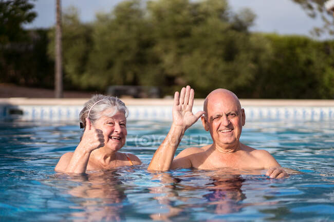 Joyeux couple de personnes âgées saluant tout en nageant dans la piscine et en regardant la caméra — Photo de stock