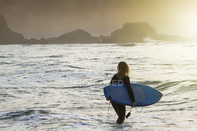 Vista posteriore di una giovane donna irriconoscibile con tavola da surf che entra in mare durante il tramonto sulla spiaggia delle Asturie, Spagna — Foto stock