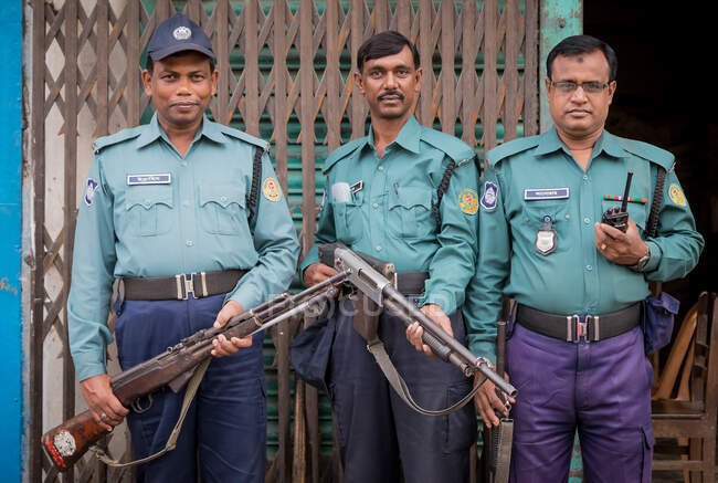 INDE, BANGLADESH - 6 DÉCEMBRE 2015 : Hommes armés ethniques en uniforme de police et casquette debout près des portes métalliques du bâtiment endommagé et regardant la caméra — Photo de stock