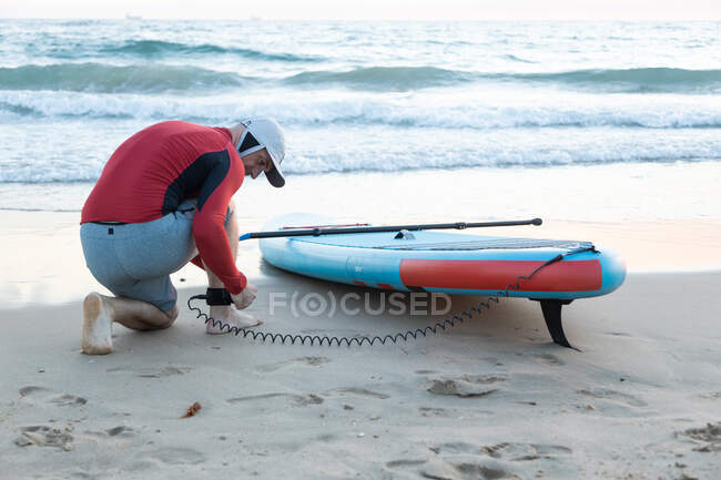 Вид сёрфера в гидрокостюме, надевающего поводок на лодыжку на доске SUP, готовящегося кататься на веслах по берегу моря — стоковое фото