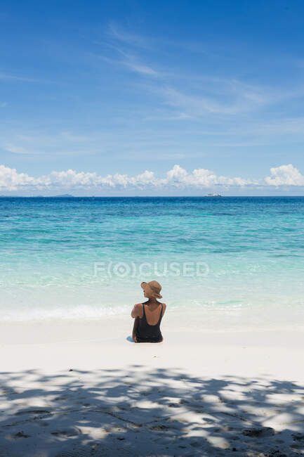 Back view female tourist in swimsuit and straw hat sitting in transparent sea during trip in Malaysia — Stock Photo