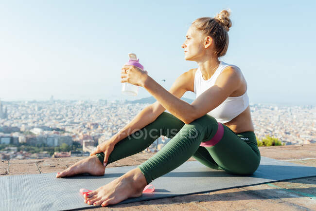 Молода споглядальна жінка-спортсменка з пляшкою води та смугою опору торкається чола, дивлячись на міський дах — стокове фото