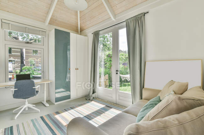 Mesa de madera con portátil situada cerca de armario blanco en habitación de luz con cómodo sofá en día soleado - foto de stock