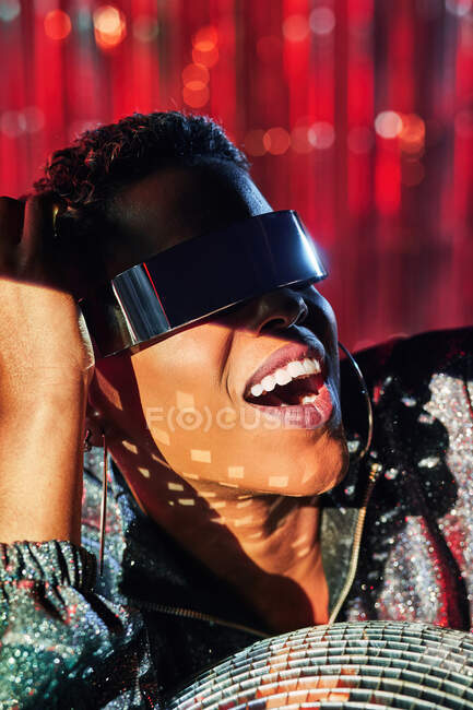 Mulher étnica feliz na moda em óculos cyberpunk e relógio de pulso tocando bola de discoteca em raio de luz — Fotografia de Stock