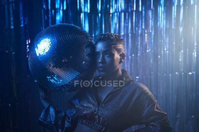 Moda joven mujer afroamericana en chaqueta de cultivo con bola de purpurina mirando hacia arriba en la luz azul en el club nocturno - foto de stock