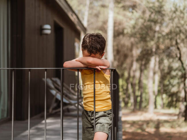 Одинокий обиженный ребенок, опирающийся на деревянную террасу дома в лесу в солнечный день — стоковое фото
