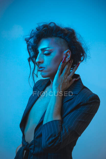 Young transsexual male model with makeup in stylish jacket looking down on blue background — Stock Photo