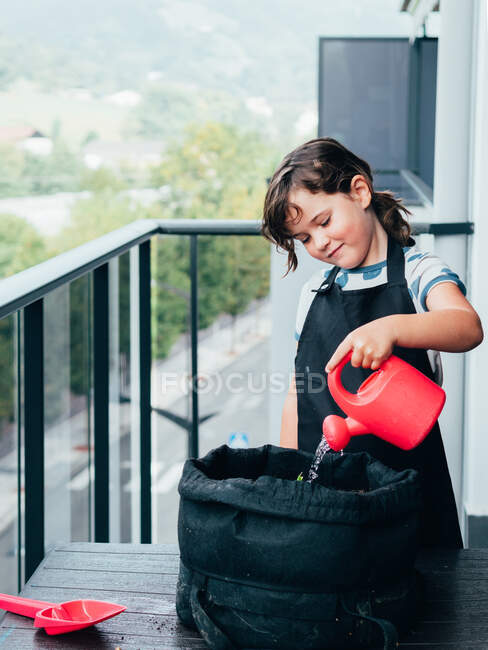 Весела маленька дівчинка з темним волоссям у фартусі стоїть і поливає рослину з горщиками на балконі вдень — стокове фото