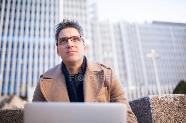 Low angle of young male in trendy outfit and eyeglasses sitting on bench and browsing netbook while working on project on street — Stock Photo