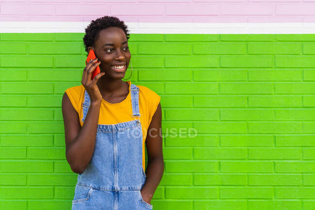 Задоволена афро-американська жінка стоїть з рукою в кишені біля яскравої стіни і розмовляє по мобільному телефону, дивлячись на — стокове фото