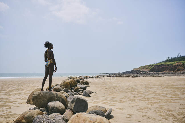 Вид сбоку на юную афроамериканку в купальнике, стоящую на валуне и отводящую взгляд в сторону — стоковое фото