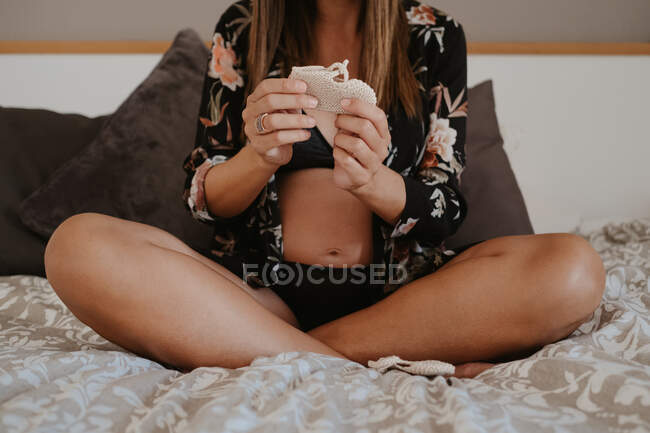 Cultiver femme enceinte méconnaissable en sous-vêtements assis avec les jambes croisées tout en regardant des chaussettes de bébé assis sur un lit doux dans la maison — Photo de stock