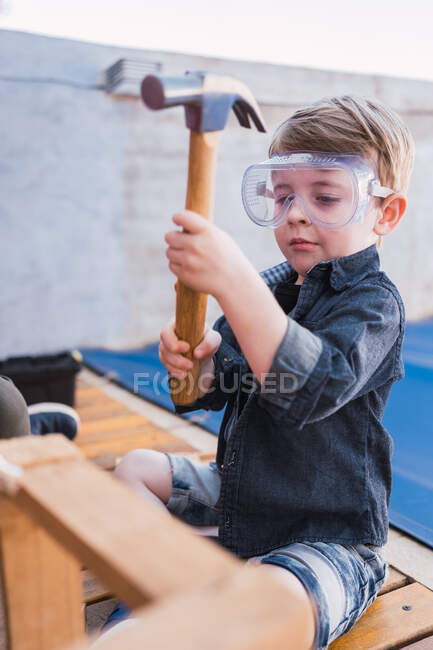 Enfant en chemise denim et lunettes en plastique assis avec marteau contre pièce en bois à la lumière du jour — Photo de stock