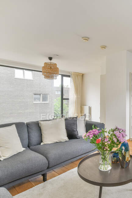 Интерьер современной гостиной с подушками на диване против цветущих цветов в вазе на столе под лампой в доме — стоковое фото