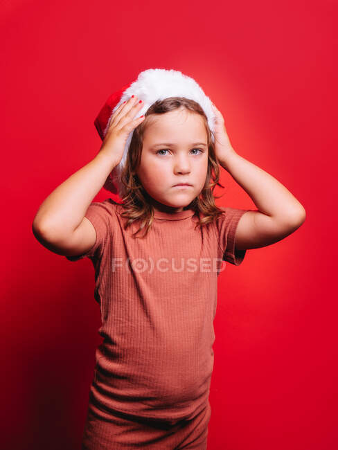 Entzückendes kleines Mädchen in lässiger Kleidung und Weihnachtsmütze, das den Kopf berührt, während es vor rotem Hintergrund steht und in die Kamera schaut — Stockfoto