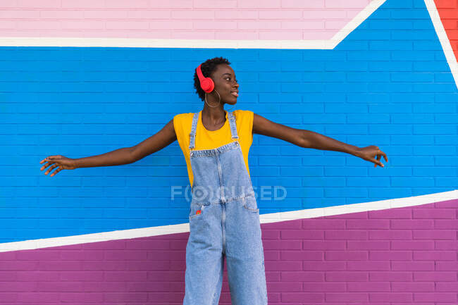 Carefree African American female listening to music in wireless headphones and outstretching arms against bright wall — Stock Photo