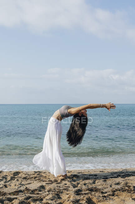 Vista laterale della femmina etnica appoggiata indietro mentre in piedi in Ashta Chandrasana posa durante la pratica dello yoga sulla costa sabbiosa contro l'oceano — Foto stock