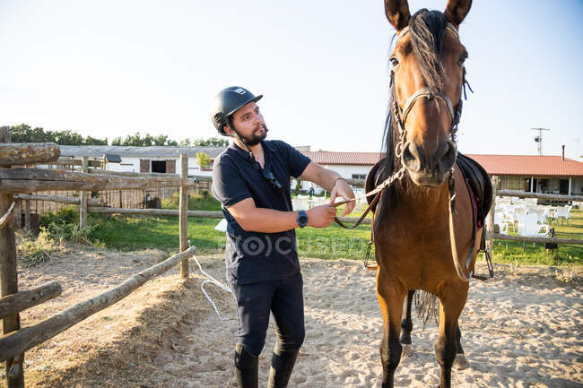 Hombre adulto en casco protector sosteniendo semental por riendas contra establos de escuela de equitación en el campo - foto de stock