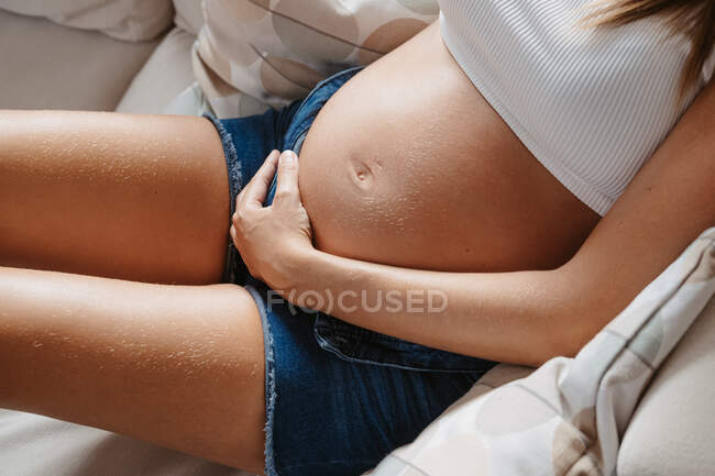 Cropped unrecognizable adult expectant female caressing belly while resting on couch at home — Stock Photo