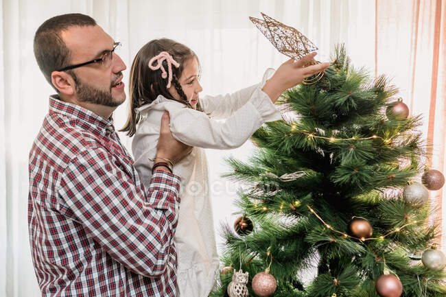 Vista lateral de padre levantando hija ayudando a decorar abeto de Navidad con estrella festiva mientras se prepara para la celebración - foto de stock
