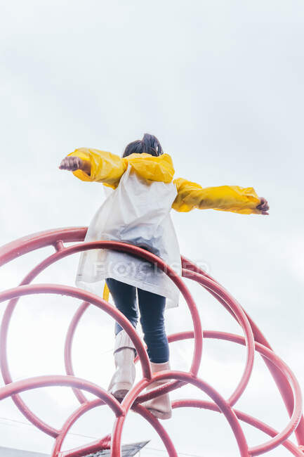 Visão traseira da criança anônima de capa de chuva em pé com os braços separados em construção simétrica sob o céu claro em tempo chuvoso — Fotografia de Stock