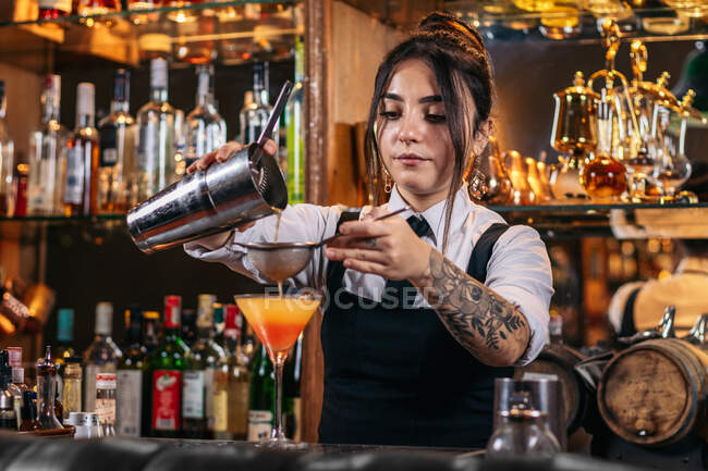 Молодая татуированная брюнетка разливает алкоголь из шейкера через решето, делая коктейль восхода текилы во время работы в баре — стоковое фото