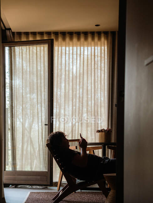 Вид сбоку на силуэт анонимного человека, сидящего в кресле и просматривающего мобильный телефон во время охлаждения в комнате дома в выходные — стоковое фото