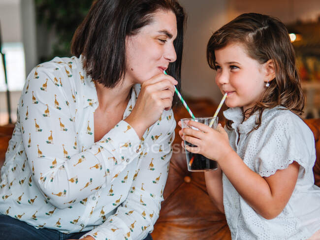 Smiling girl sharing glass of sweet soda with mother drinking with straw in cafe — Stock Photo