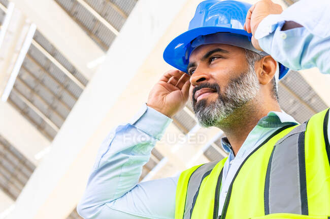 D'en bas du superviseur masculin ethnique mature en gilet portant un casque tout en regardant loin debout près de la centrale solaire — Photo de stock