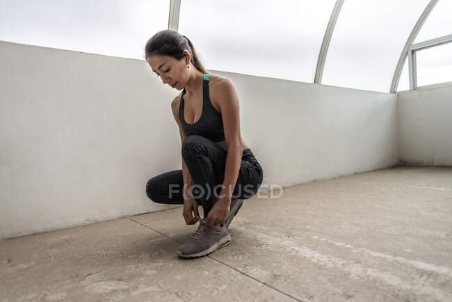 Young ethnic female athlete in sports clothes tying shoelace on footwear while squatting on floor before training — Stock Photo