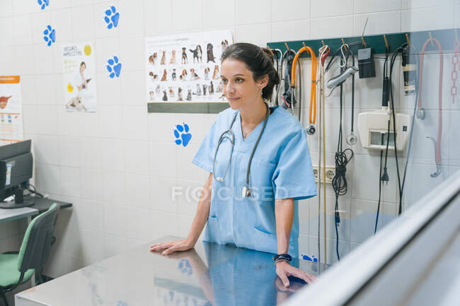 Дружелюбная женщина-ветеринар в медицинской форме со стетоскопом смотрит вперед за металлическим столом в больнице — стоковое фото