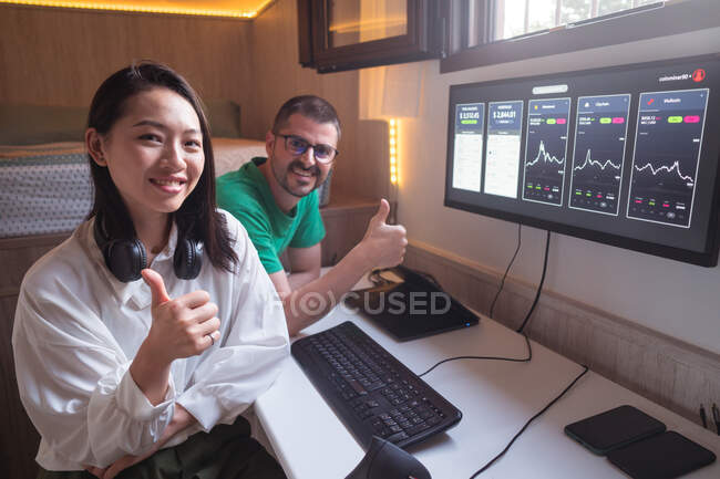 Optimistic diverse cryptocurrency traders showing thumb ups while analyzing graphs of cryptocurrency market and looking at camera — Stock Photo