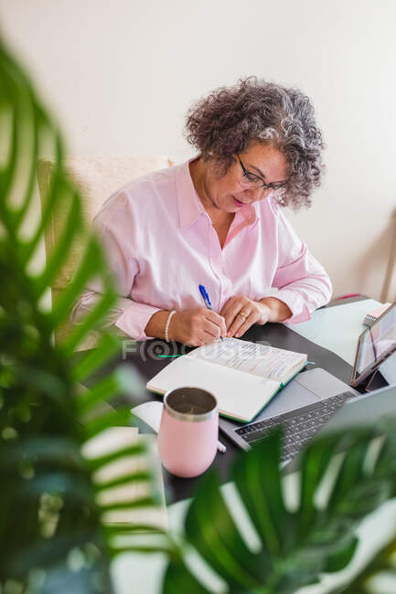 Alto ángulo de emprendedora senior tomando notas en la agenda mientras trabaja en el escritorio con netbook en el espacio de trabajo - foto de stock